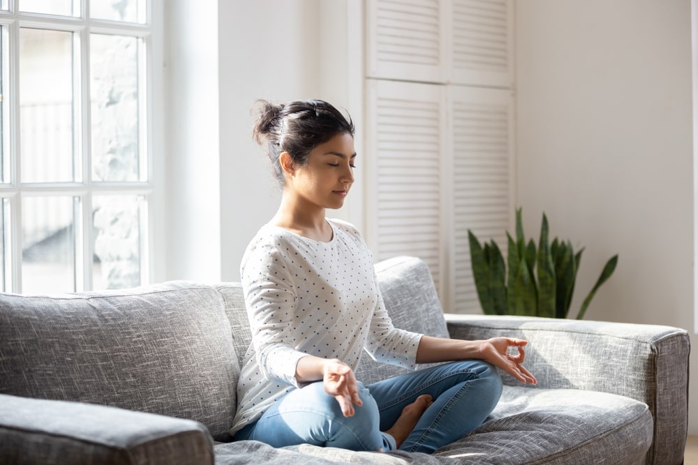 mulher meditando sentada no sofá para ilustrar texto sobre benefícios da meditação
