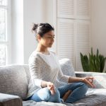 mulher meditando sentada no sofá para ilustrar texto sobre benefícios da meditação