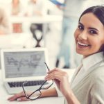 mulher sorrindo com óculos na mão na frente de um notebook para ilustrar texto sobre período de experiência no trabalho