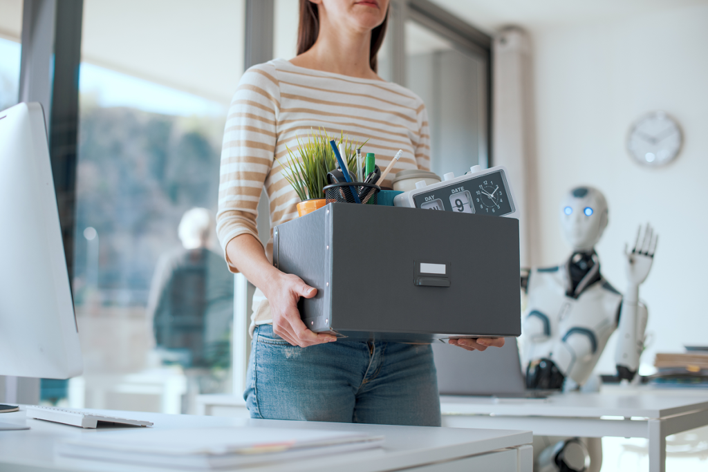 Mulher saindo do escritório carregando uma caixa com seus pertences. Robô abana ao fundo. Inteligência artificial e mercado de trabalho